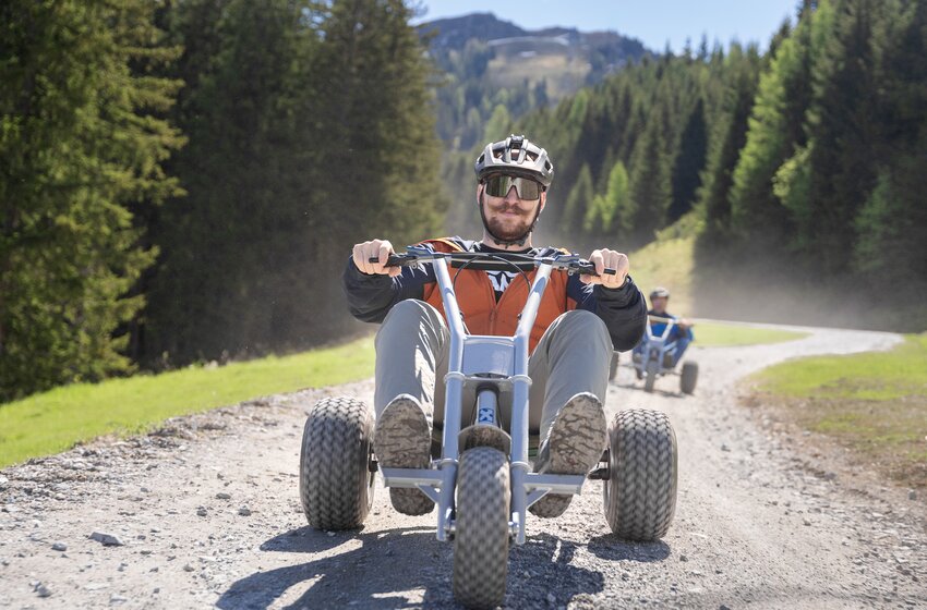 A man rides a mountain cart down a curving gravel path surrounded by forest and grass under bright sunshine | © Dorfgasteiner Bergbahnen AG