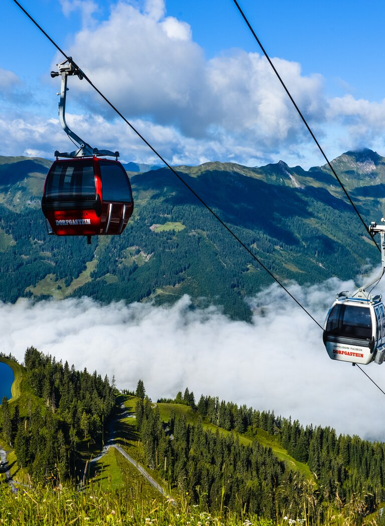 Two cable cars over Dorfgastein with forest, pond and clouds below, set against an alpine mountain backdrop. | © Dorfgasteiner Bergbahnen AG