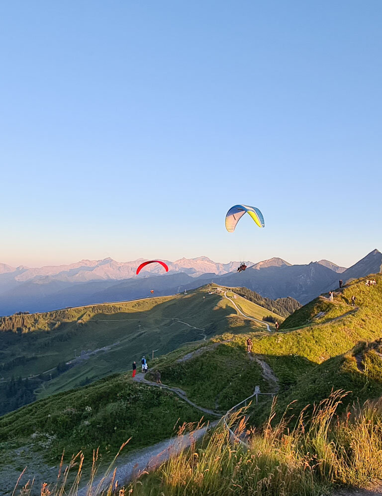 Two paragliders float above green Alps at sunset, with hikers and a mountain station in the foreground. | © Dorfgasteiner Bergbahnen AG