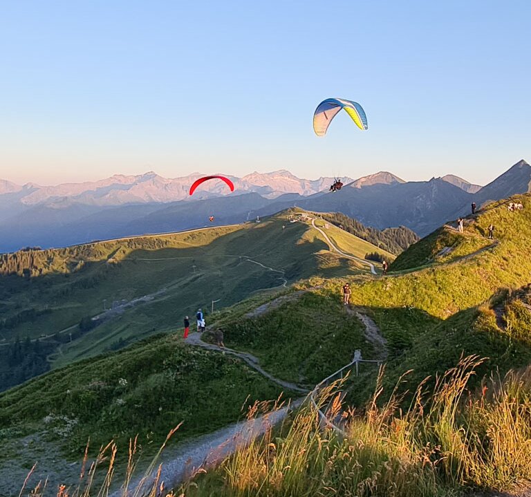 Two paragliders float above green Alps at sunset, with hikers and a mountain station in the foreground. | © Dorfgasteiner Bergbahnen AG