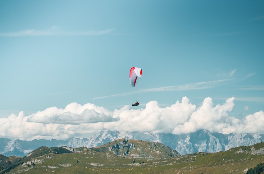 A single paraglider soars above green mountains under sunlight, with clouds and the High Tauern mountain range in the background. | © Dorfgasteiner Bergbahnen AG