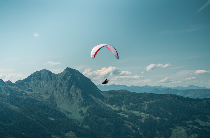 A paraglider floats in the sun before a steep green mountain ridge, overlooking forested valleys and distant alpine peaks. | © Dorfgasteiner Bergbahnen AG