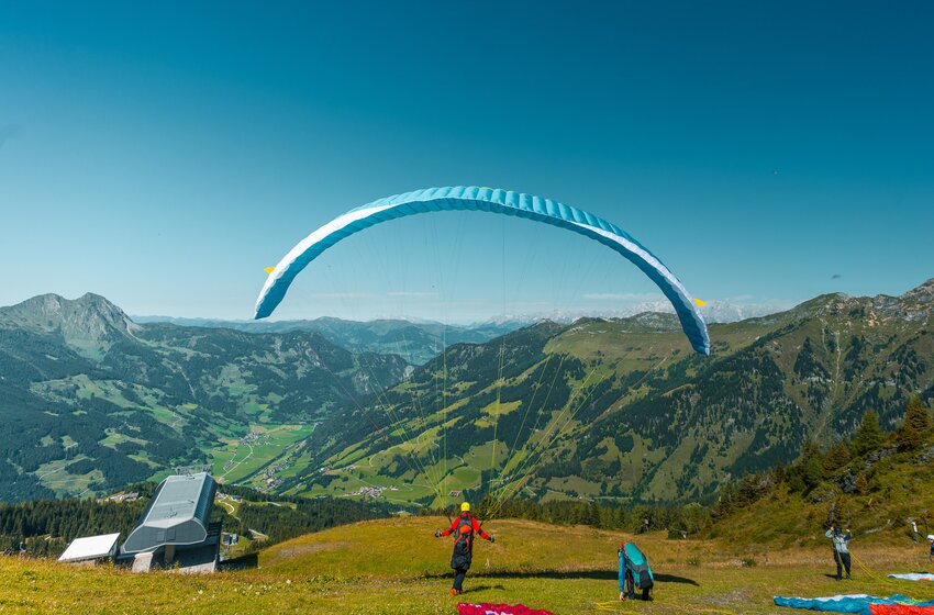 Ein Paragleiter startet mit hellblauem Schirm oberhalb der Bergstation, mit Blick auf das grüne Gasteinertal und umliegende Berge. | © Dorfgasteiner Bergbahnen AG