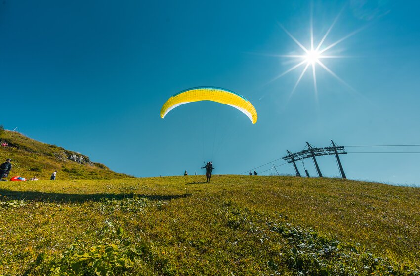 Ein Paragleiter hebt auf einer sonnigen Wiese mit gelbem Schirm ab, während Zuschauer am Hang und eine Seilbahn im Bild sind. | © Dorfgasteiner Bergbahnen AG