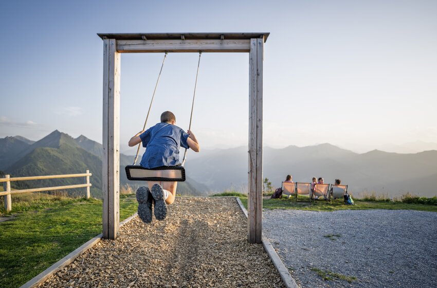 Kind schaukelt auf Holzschaukel auf einem Berg mit Blick auf Alpen, während Menschen im Hintergrund sitzen | © Dorfgasteiner Bergbahnen AG