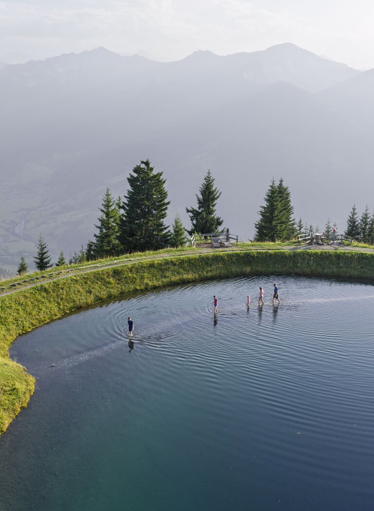 Menschen stehen in einem Bergsee auf einer Anhöhe, umgeben von Wiesen, Tannen und Bergpanorama. | © Dorfgasteiner Bergbahnen AG