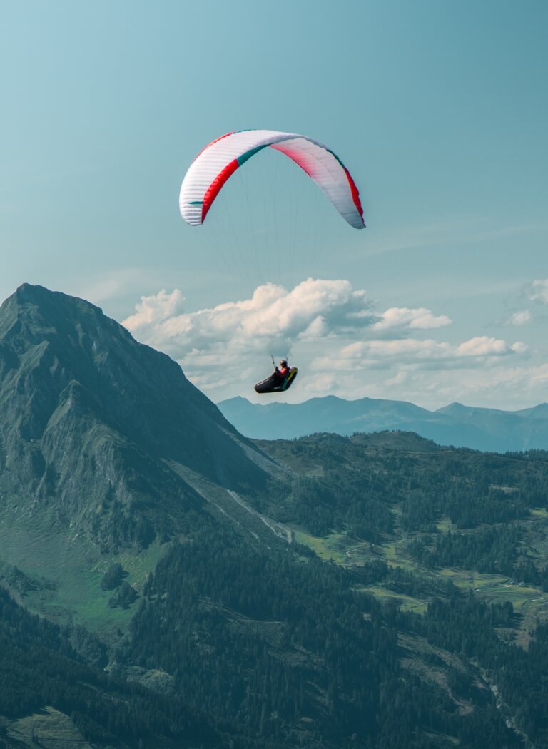 Gleitschirmflieger schwebt über bewaldeten Bergen und grünen Tälern unter blauem Himmel mit Wolken. | © Dorfgasteiner Bergbahnen AG