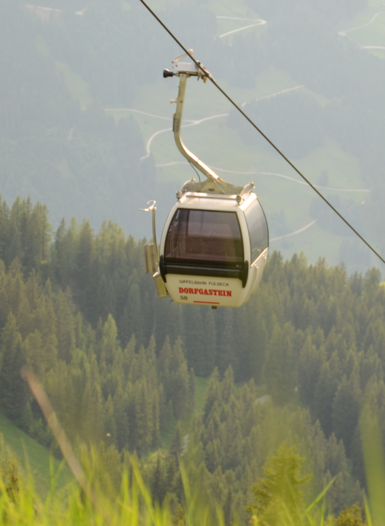 Rote und weiße Gondeln der Gipfelbahn Fulseck schweben über grünen Almwiesen vor bewaldeten Berghängen. | © Dorfgasteiner Bergbahnen AG