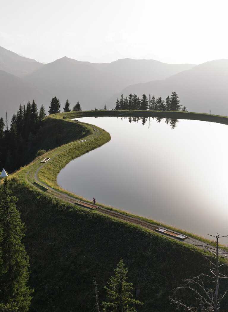 Spiegelsee auf einem Bergkamm mit ruhiger Wasseroberfläche, Wanderweg, Nadelbäumen und Bergkulisse im Abendlicht. | © Dorfgasteiner Bergbahnen AG