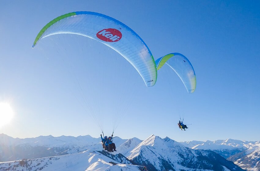 Two tandem paragliders fly in sunny weather above snowy Alpine landscape and blue sky. | © Dorfgasteiner Bergbahnen AG