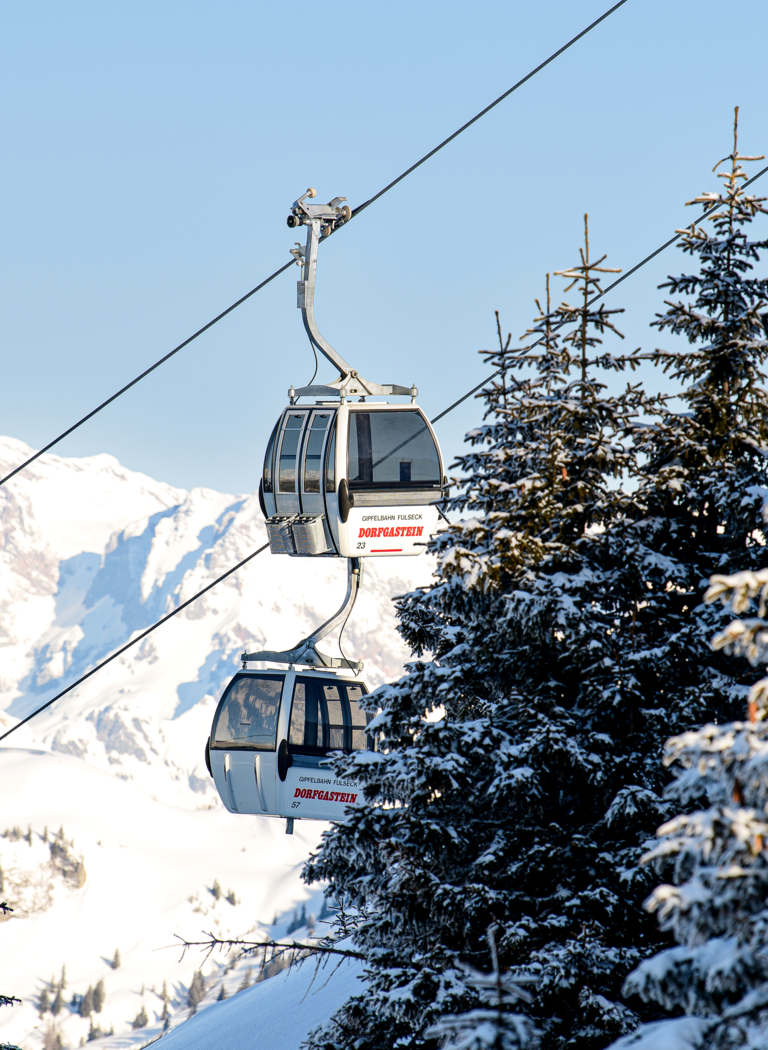Two cable car cabins travel between snow-covered trees with alpine mountains in the background under clear sky | © Dorfgasteiner Bergbahnen AG