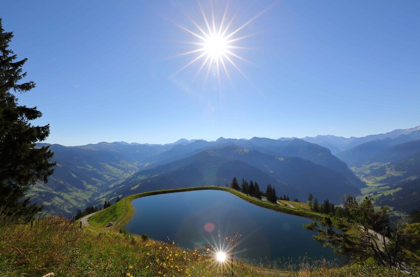 Speichersee im Großarltal mit Sonnenreflexion und weitem Blick ins Tal | © Grossarler Bergbahnen GmbH & Co KG Bergsee mit Spiegelung der Sonne oberhalb des Großarltals, mit Blick auf grüne Hügel und entfernte Gipfel | © Grossarler Bergbahnen GmbH & Co KG