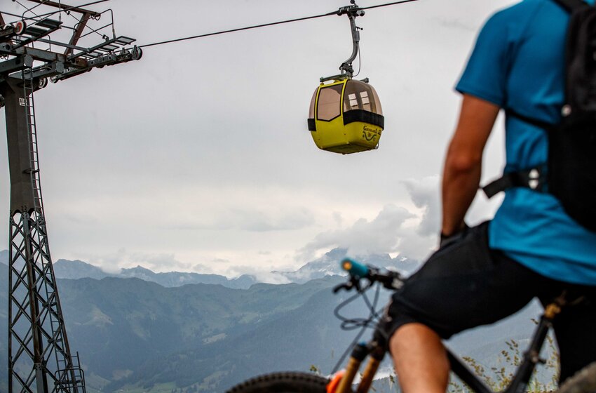 Mountain biker from behind facing yellow retro gondola of Großarl cable car with cloudy sky and alpine scenery | © Grossarler Bergbahnen GmbH & Co KG