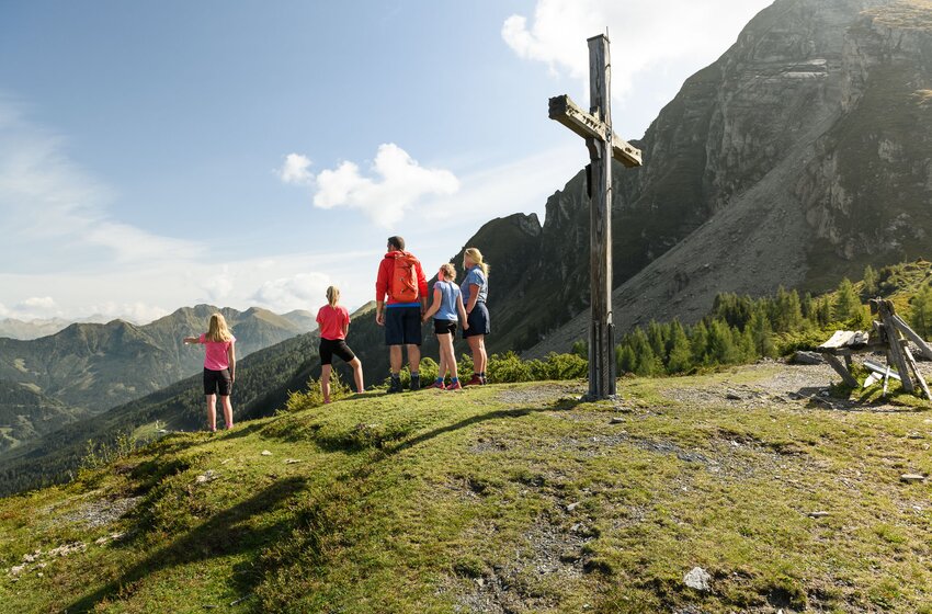 Eine Familie steht am Hahnbalzköpfl beim Gipfelkreuz und blickt ins Gasteinertal und auf die umliegenden Berge. | © Gasteiner Bergbahnen AG