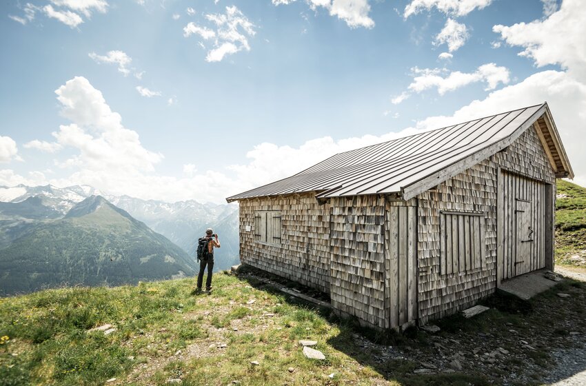 Wanderer vor rustikaler Holzhütte am Stubnerkogel mit Ausblick auf grüne Berge und die Hohen Tauern. | © Gasteiner Bergbahnen AG