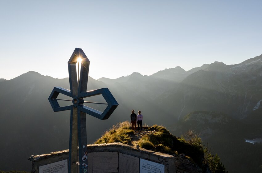 Zwei Wanderer stehen am Gipfelkreuz auf einem Berggrat, Sonne scheint durch Metallkreuz, Alpen im Hintergrund | © Gasteiner Bergbahnen AG