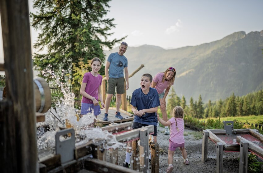 Family with three children plays at wooden water play station, water splashing, mountains and trees in background | © Dorfgasteiner Bergbahnen AG