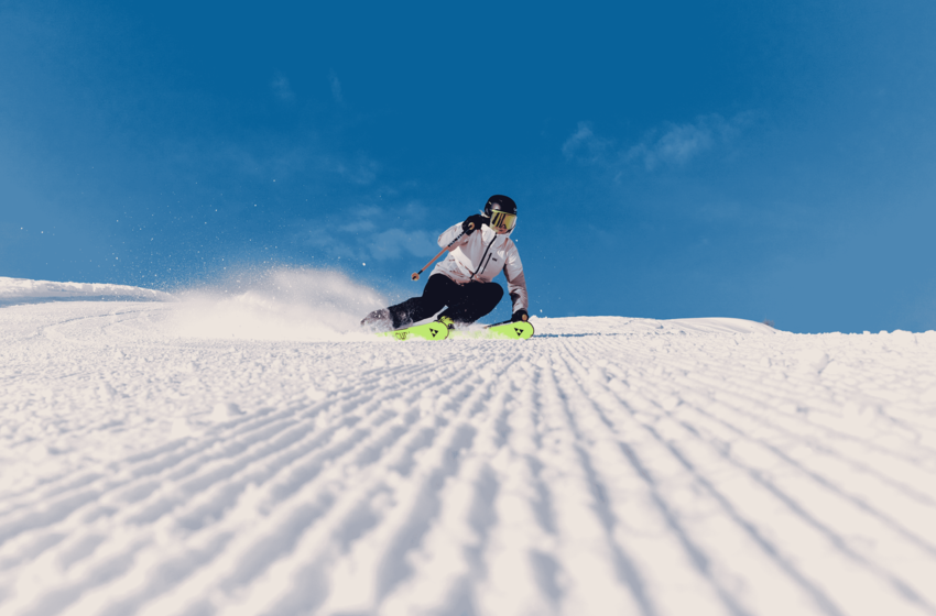 Skier in a white jacket carves a turn on a groomed piste, spraying snow beneath a clear blue sky | © Gasteiner Bergbahnen AG 