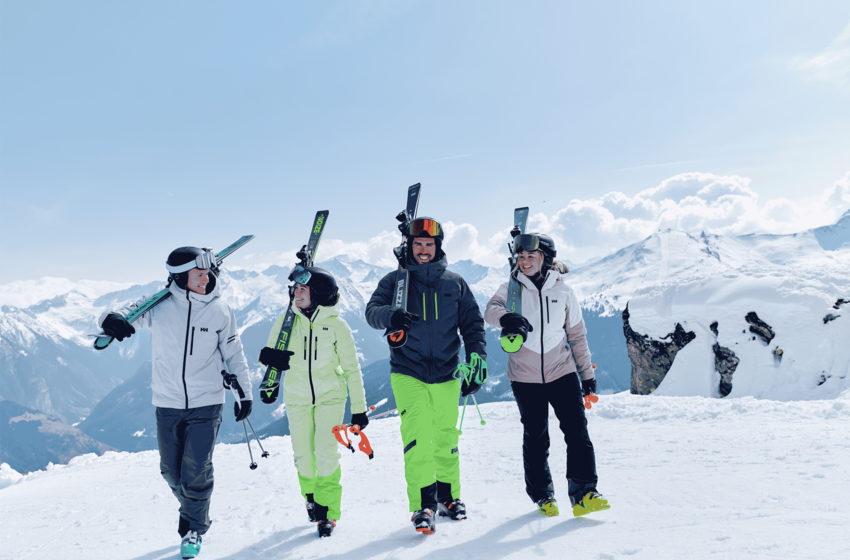 Four skiers walk across a snowy piste carrying skis and poles, with snow-covered mountain peaks behind them. | © Gasteiner Bergbahnen AG 