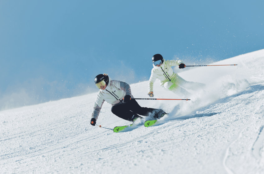 Two skiers carve tight turns on a snowy piste, spraying snow into the air beneath a clear blue sky. | © Gasteiner Bergbahnen AG 